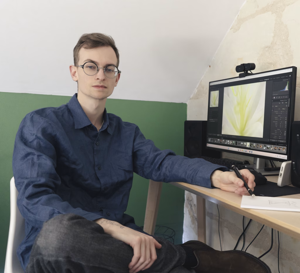un Homme assis devant un ordinateur où l'application Lightroom est ouverte, il est photographe spécialisé en retouche photo, il porte une chemise bleu foncé, il a des lunettes.